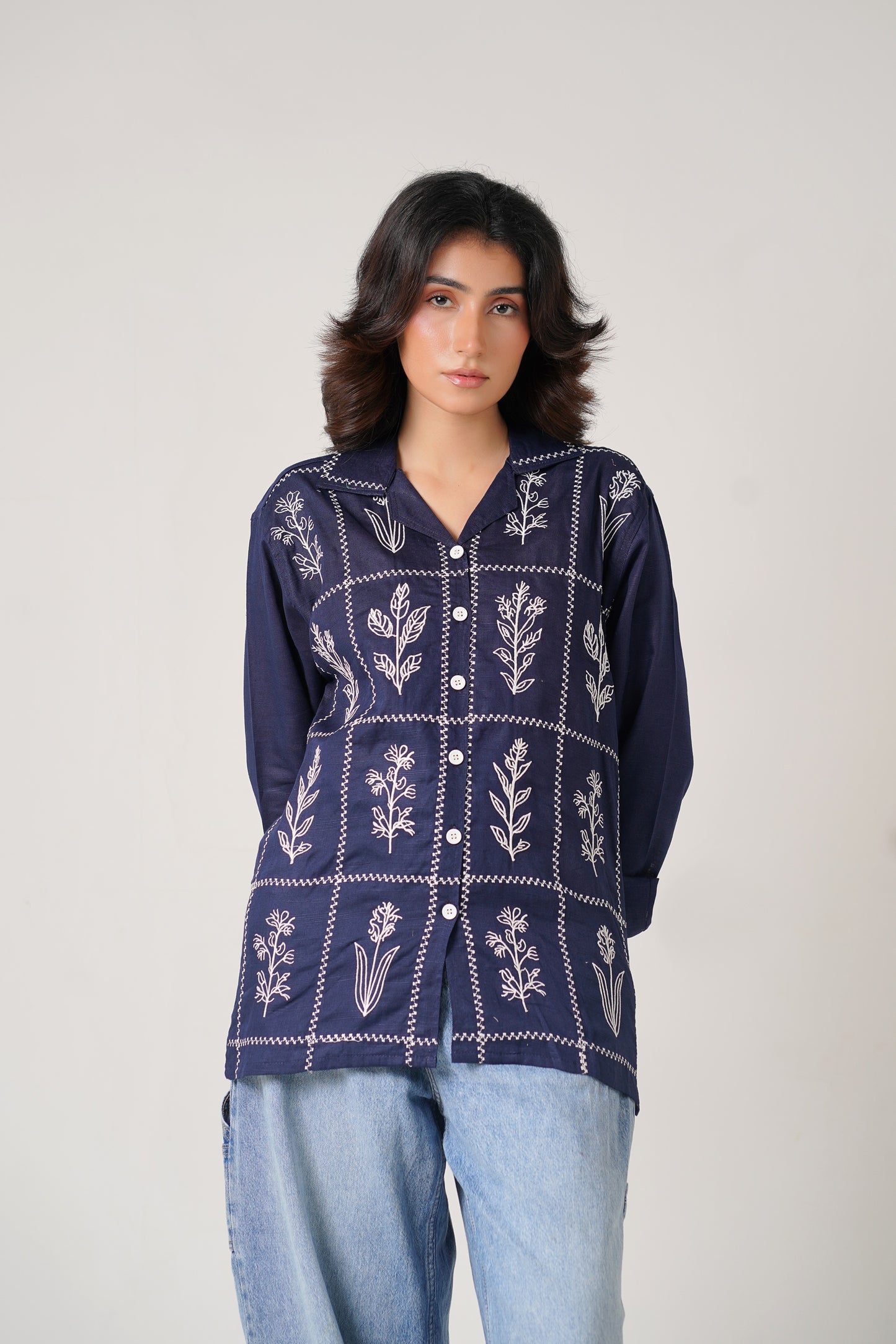 Woman wearing a navy blue embroidered shirt with light blue jeans on a plain background