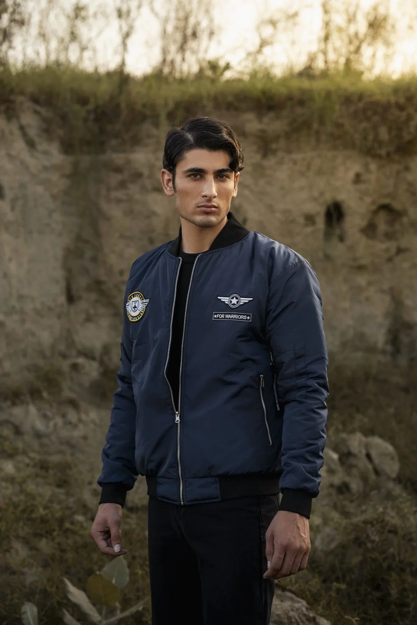 Man wearing a blue bomber jacket with logos standing in front of a natural background
