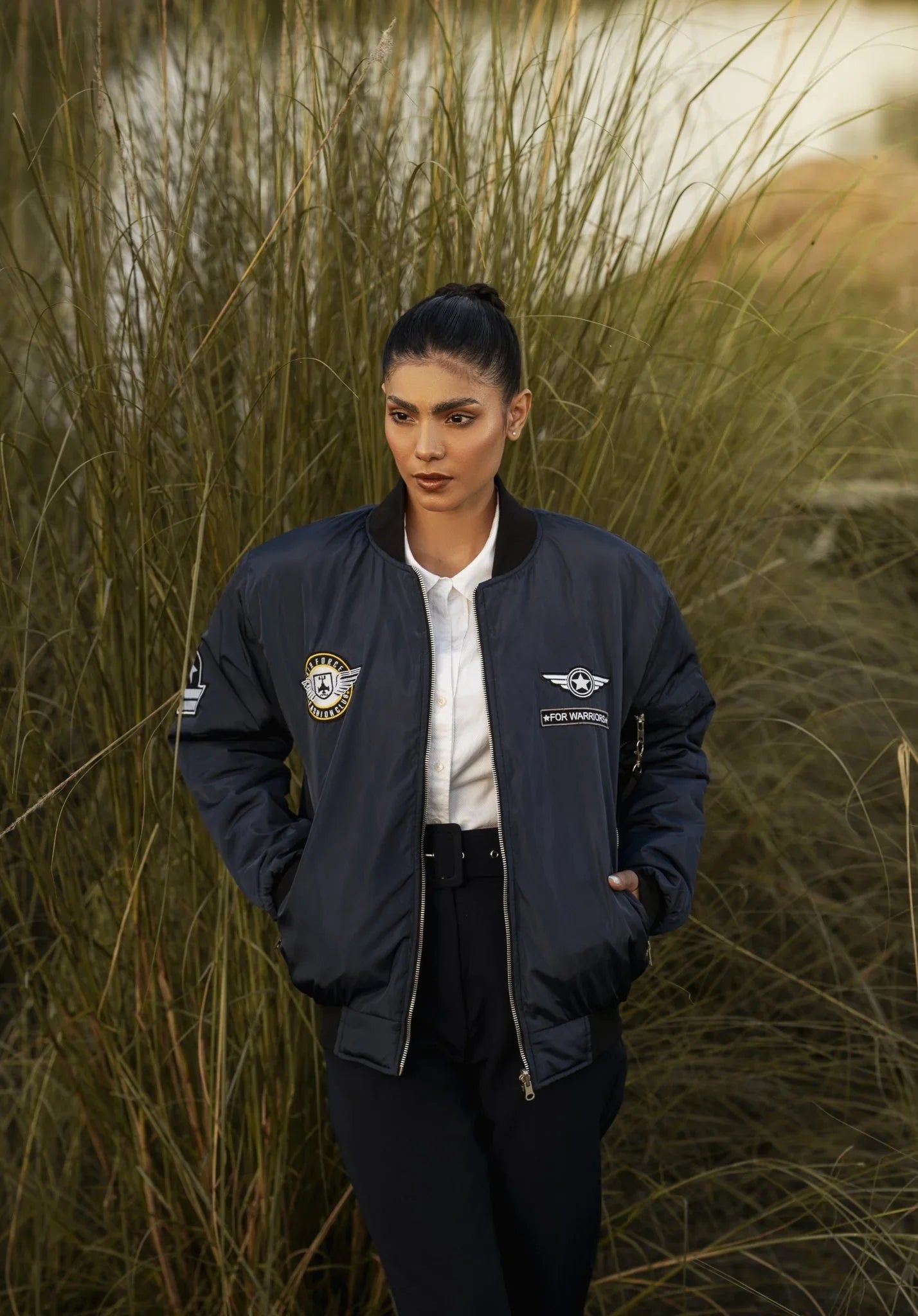 Person wearing a dark blue bomber jacket with logos, standing in tall grass.