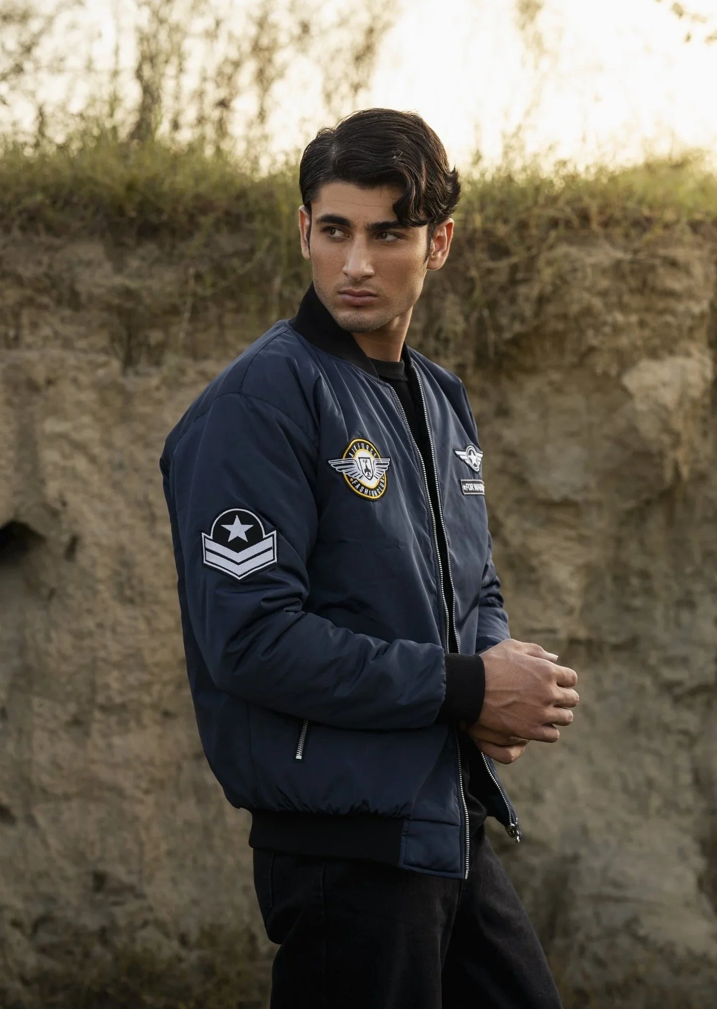 Man wearing a navy bomber jacket with a star patch against a stone wall.
