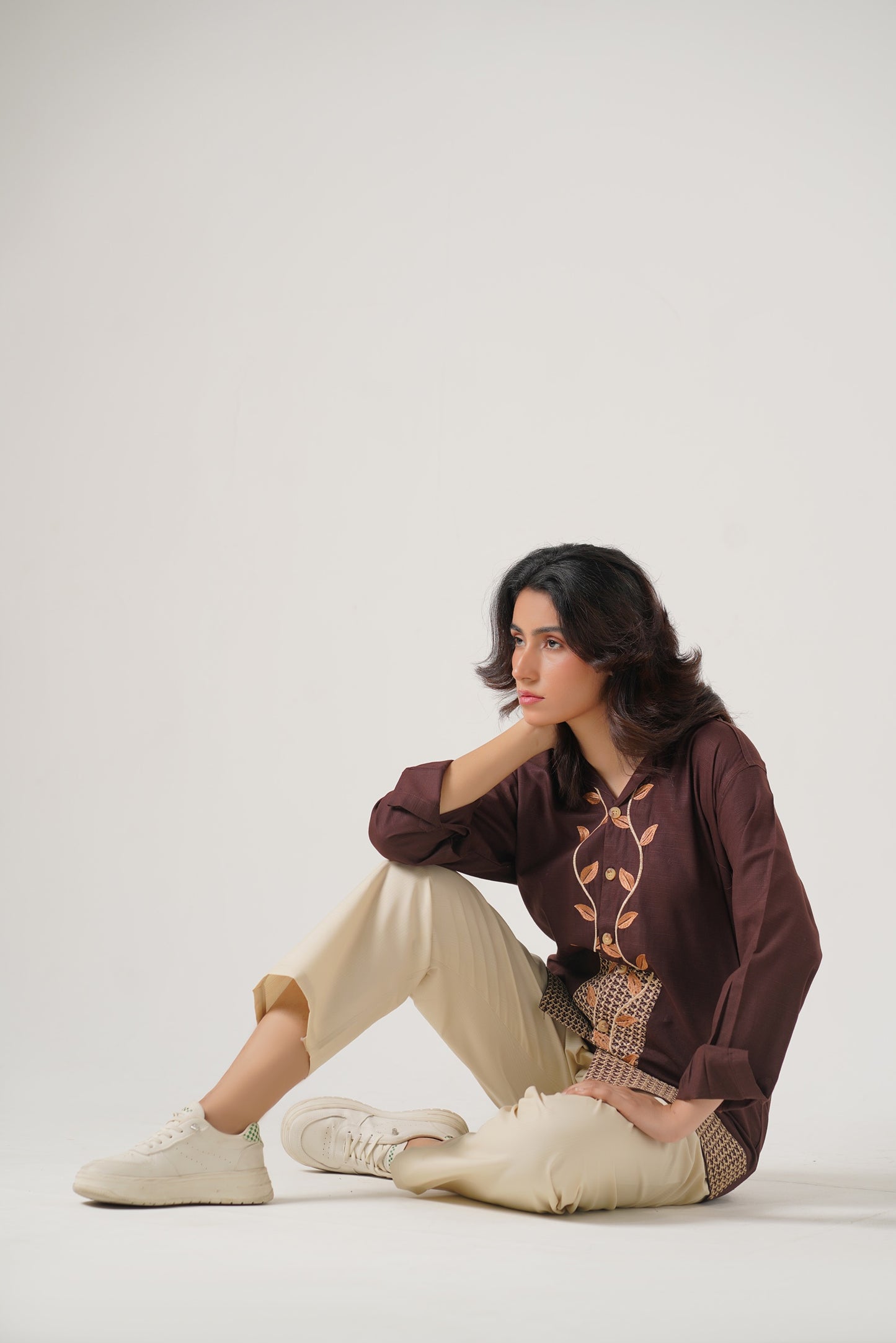 Woman sitting on a white floor wearing a brown sweater with a graphic design and beige pants.