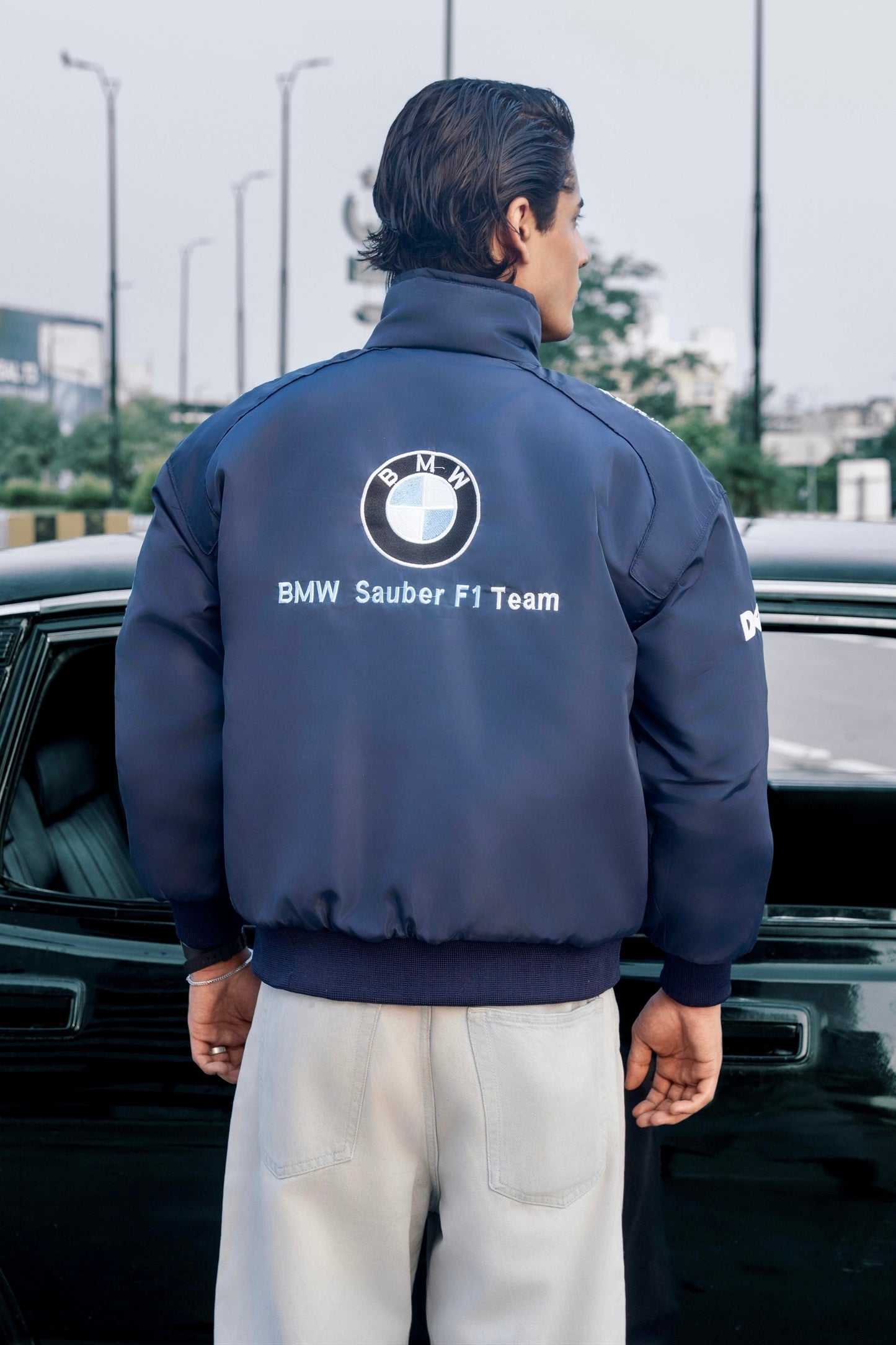 Koti Apparels navy blue BMW Sauber F1 Team racing jacket with embroidered logo worn by a man standing by a car