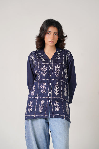 Woman wearing a navy blue embroidered shirt with light blue jeans on a plain background