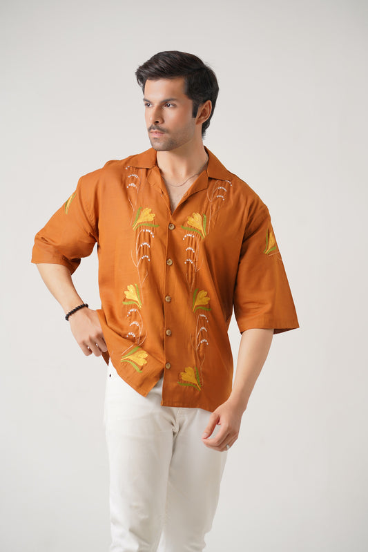 Man wearing an orange embroidered shirt with white pants on a light gray background