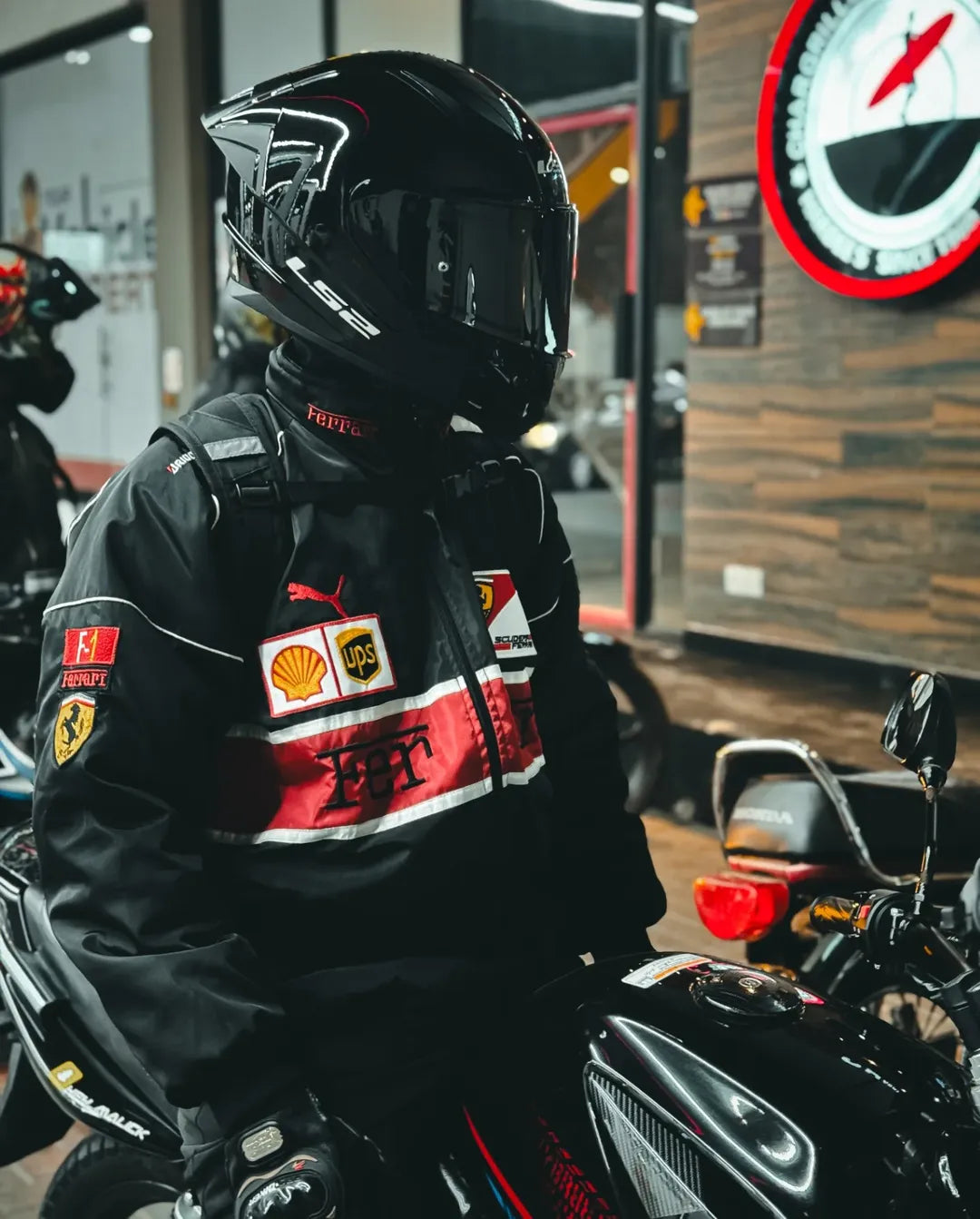Person wearing a black motorcycle helmet and ferrari jacket with various logos in an indoor setting.
