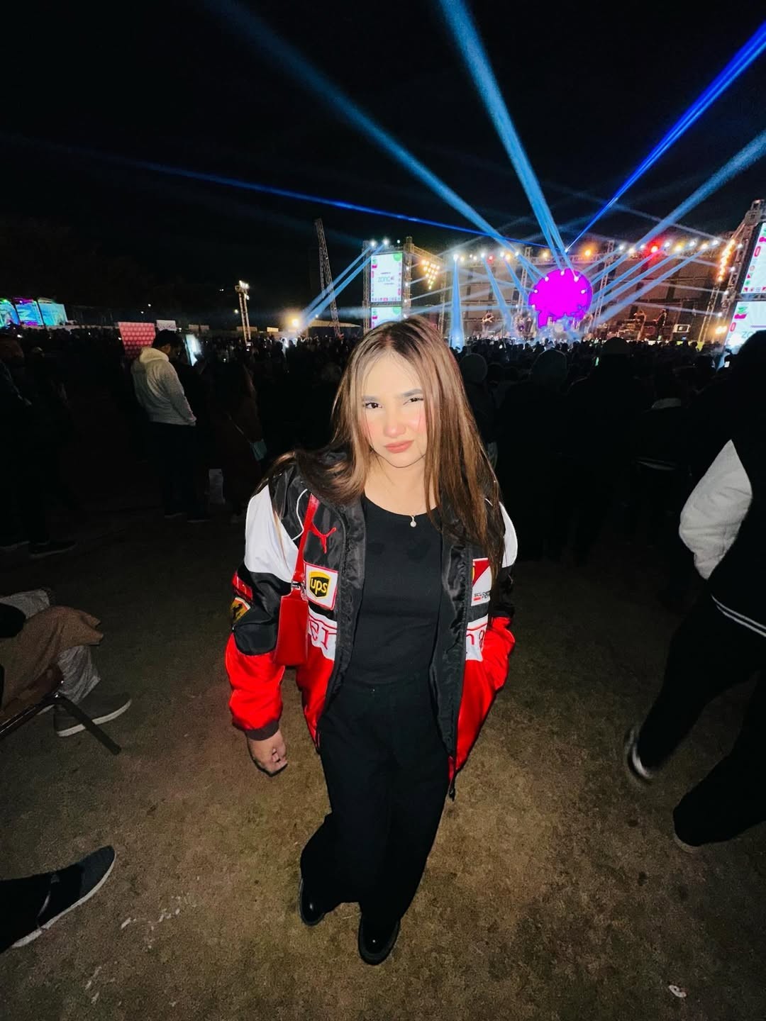 Koti Apparels red, black, and white Formula 1 racing jacket with embroidered logos, worn by a young woman at an outdoor concert.