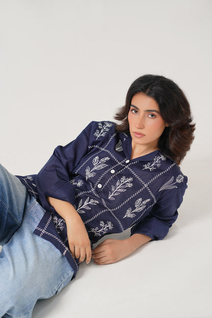 Person wearing a navy blue embroidered shirt and light blue jeans on a white background
