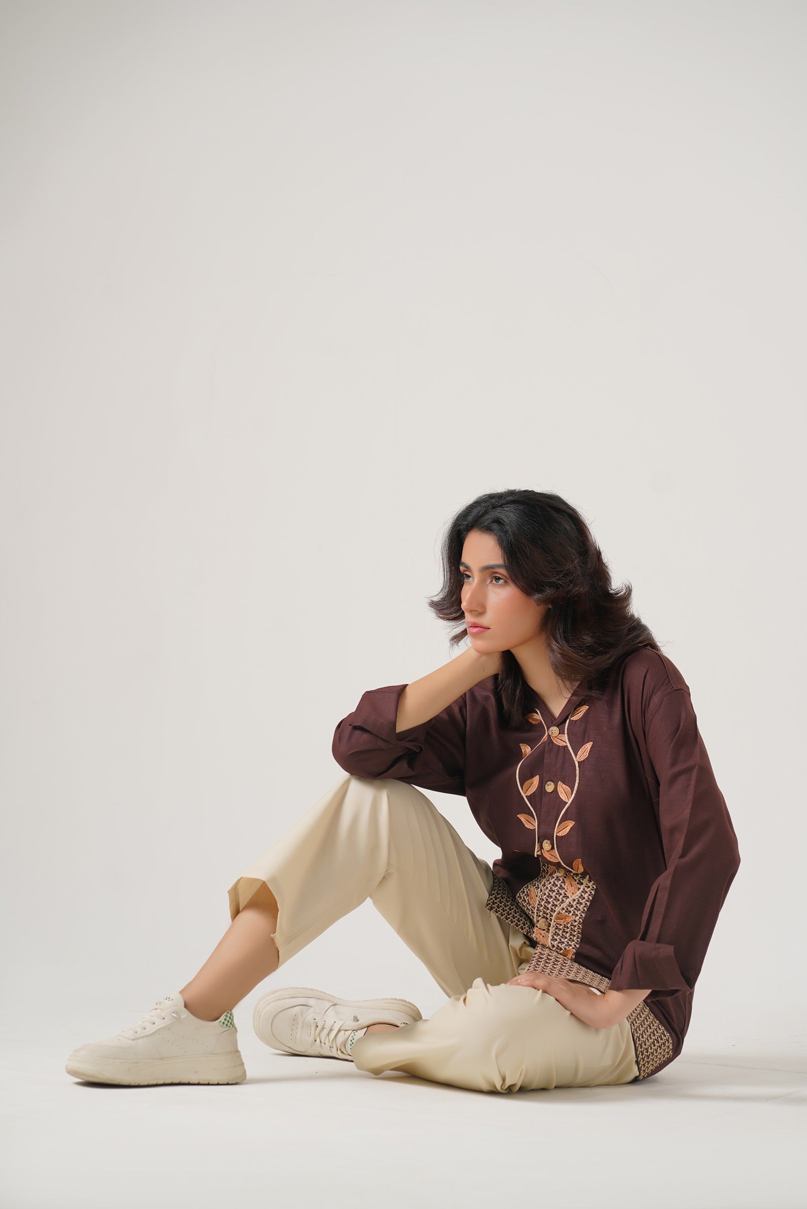 Woman sitting on a white floor wearing a brown sweater with a graphic design and beige pants.