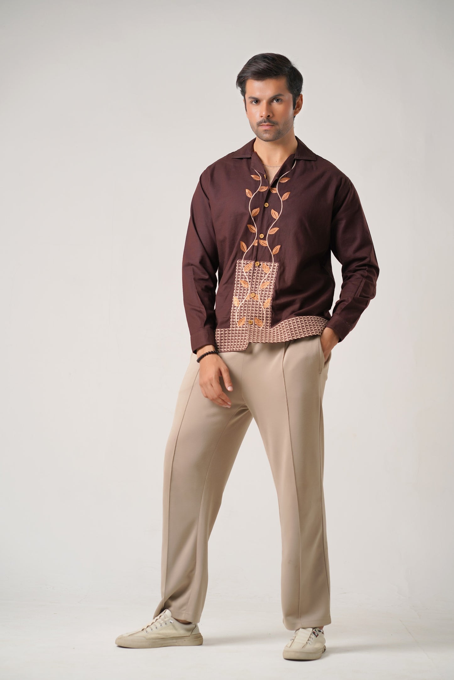 Man wearing a brown shirt with intricate designs and beige pants on a white background