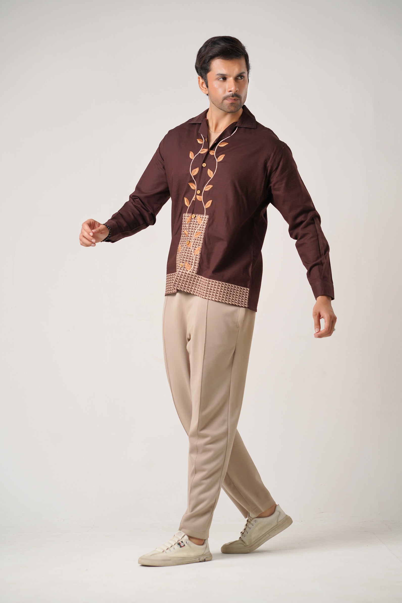 Man wearing a brown traditional shirt with intricate designs and beige pants on a white background