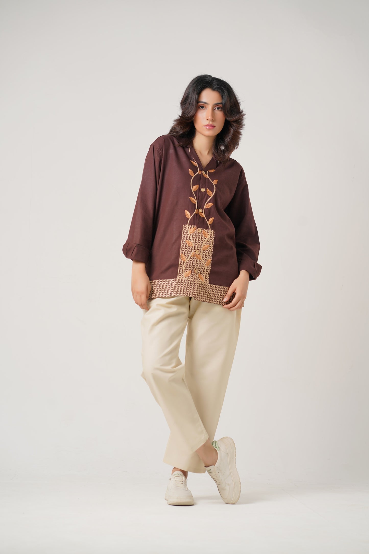 Woman wearing a brown blouse with intricate patterns and beige pants on a white background