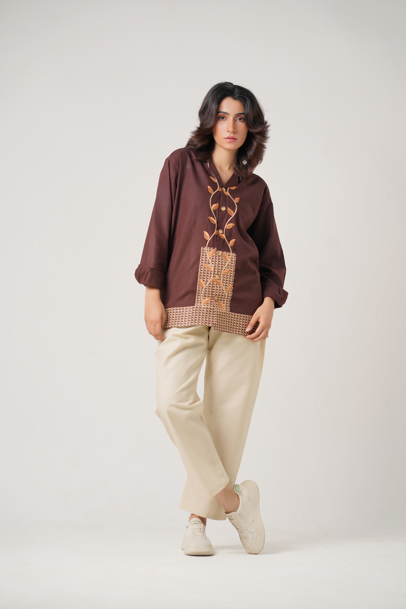 Woman wearing a brown blouse with intricate patterns and beige pants on a white background