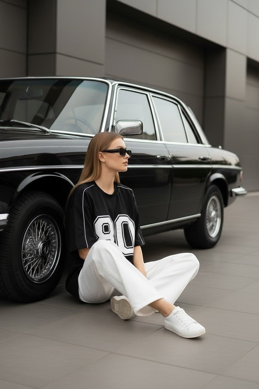 Koti Apparels black graphic baseball jersey t-shirt with white '80' graphic and piping. Model wears white pants and sneakers.