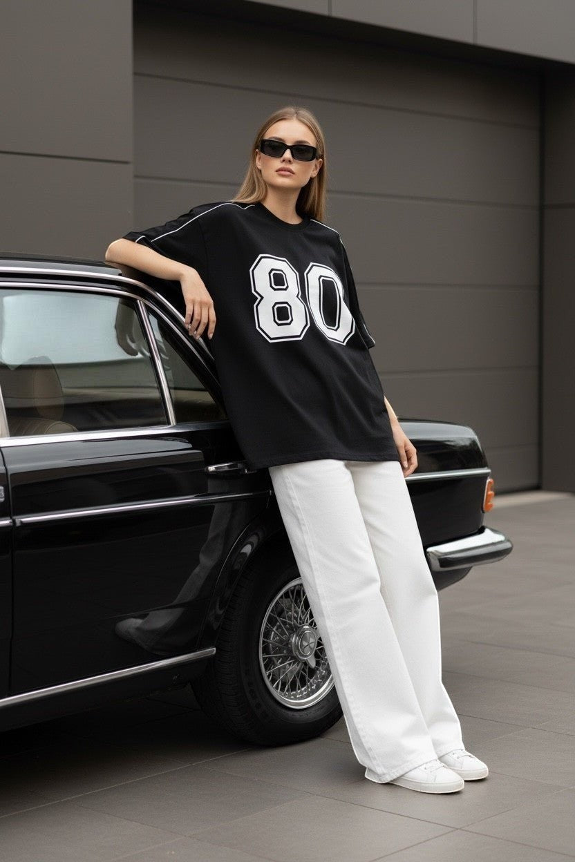 Koti Apparels black graphic baseball jersey t-shirt with white '80' graphic and piping. Model wears white pants and sunglasses.