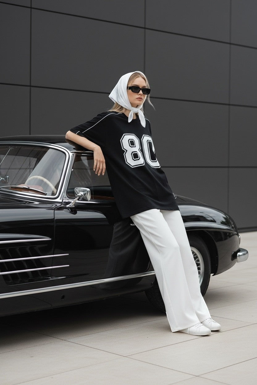 Koti Apparels black graphic baseball jersey t-shirt with white '80' graphic and piping, worn by a woman leaning on a vintage car.