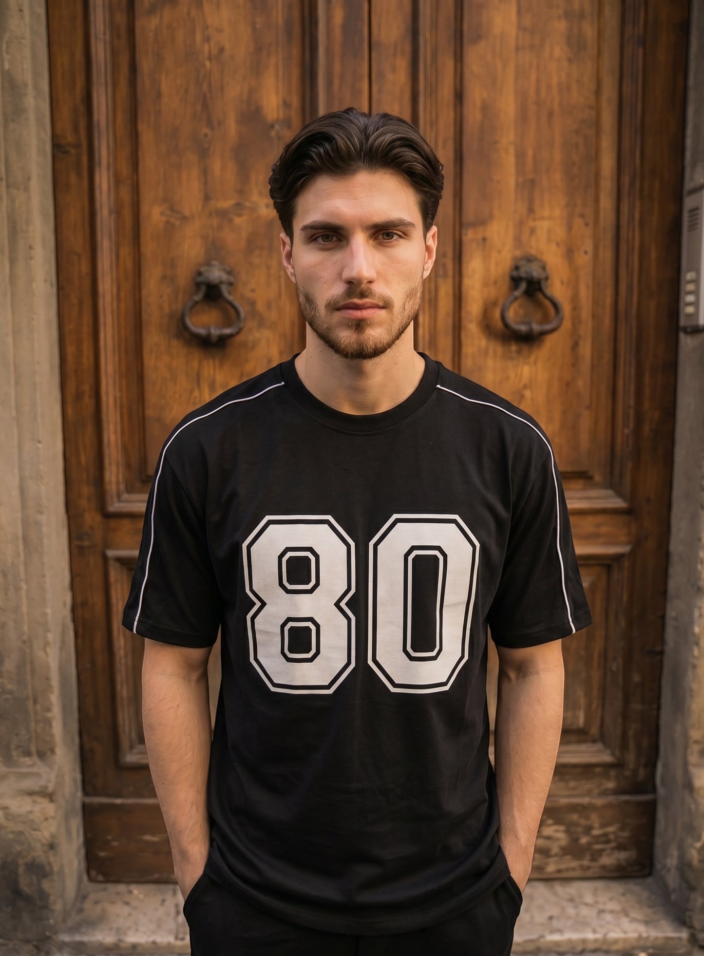 Koti Apparels black graphic baseball jersey with white 80 print and shoulder piping worn by a man against a wood door