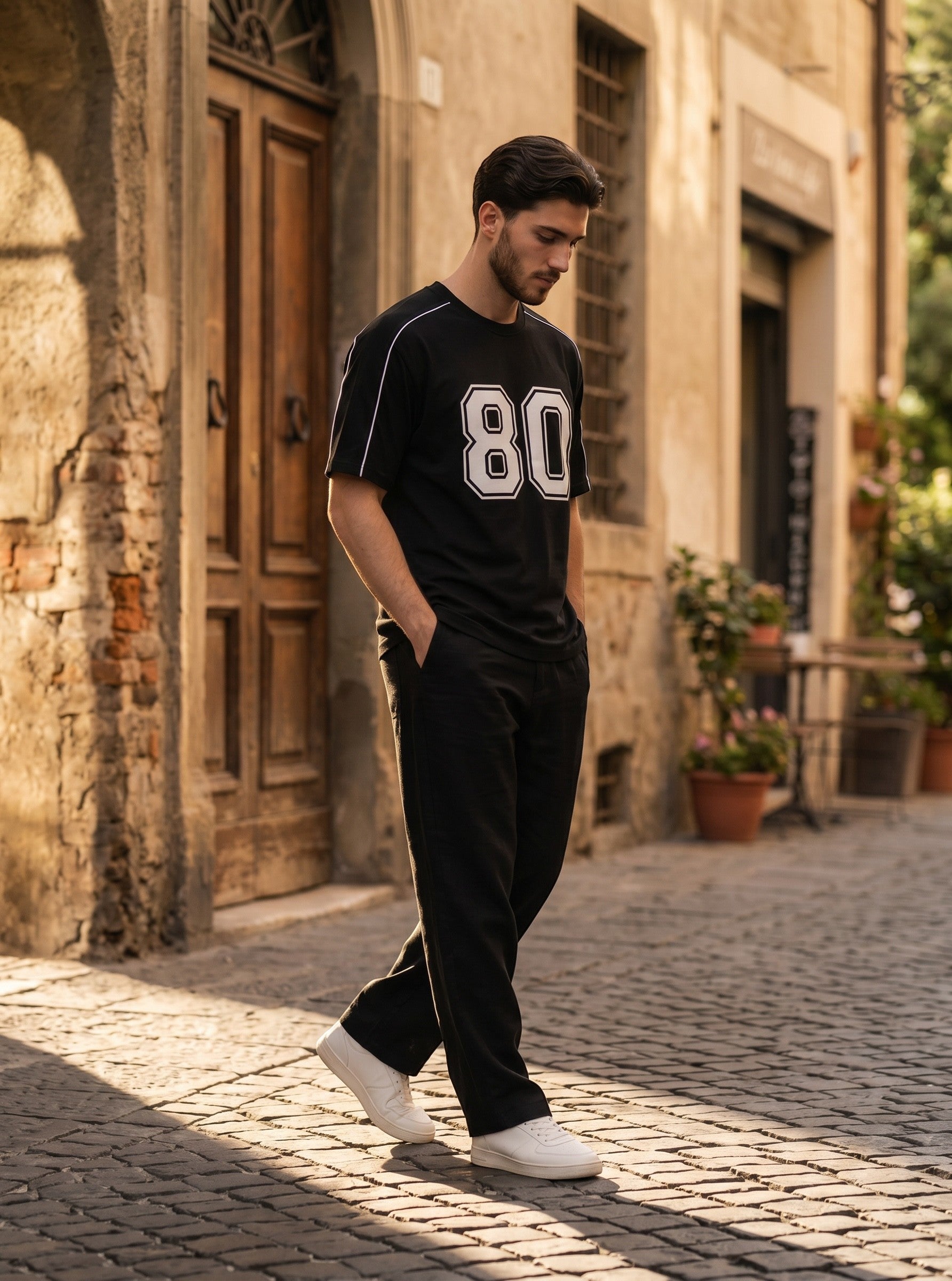 Man wearing a Koti Apparels black graphic baseball jersey with white 80 print and shoulder piping on a cobblestone street