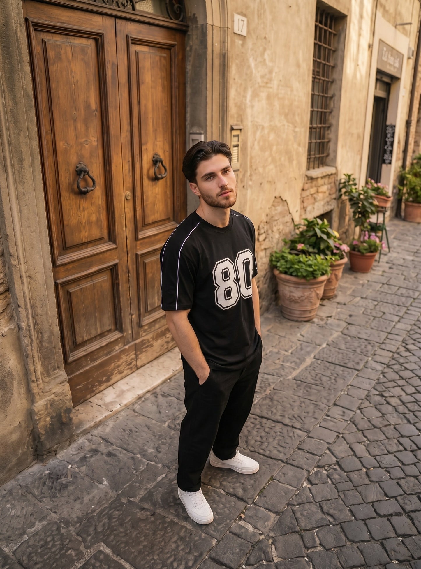 Man wearing a Koti Apparels black graphic baseball jersey with white 80 print and shoulder piping on a cobblestone street
