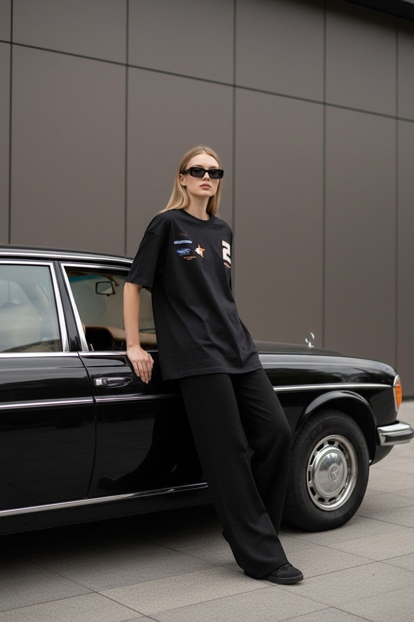 Koti Apparels black oversized cotton t-shirt with graphic print and racing patches, worn by a woman leaning on a vintage car.