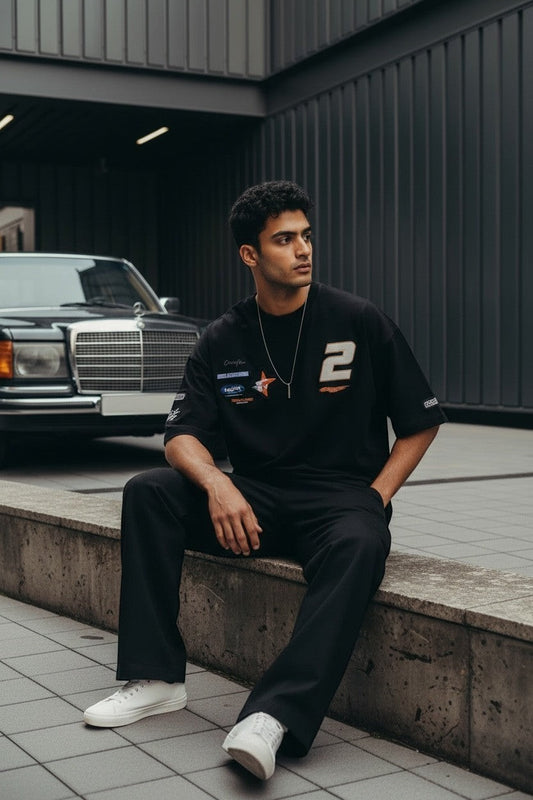 Koti Apparels black oversized cotton t-shirt with graphic print, racing patches, and number 2. Model wears it with black pants and white sneakers.