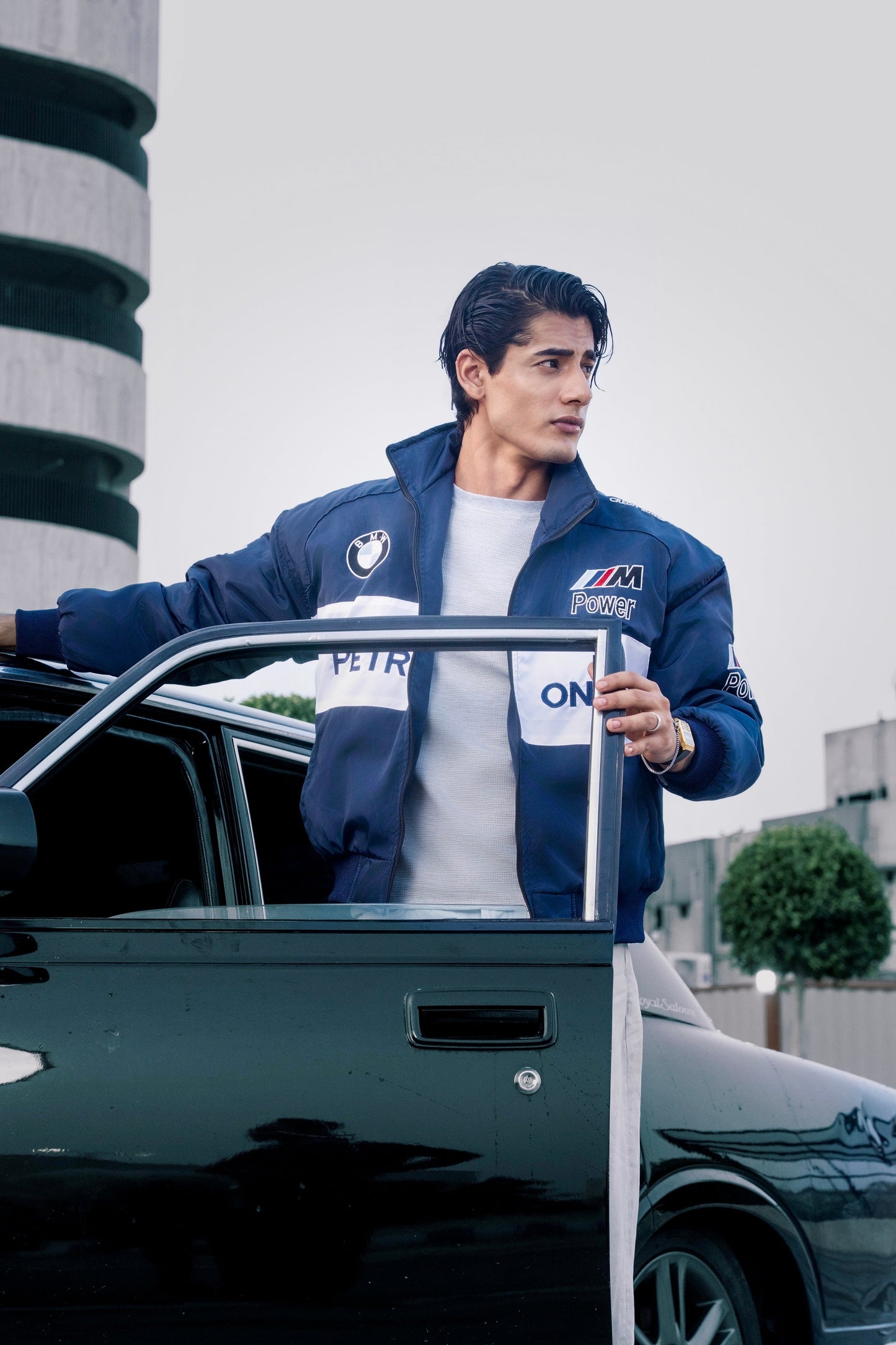 A man wearing a Koti Apparels navy blue BMW racing jacket with embroidered motorsport logos, standing by a car.