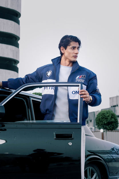 A man wearing a Koti Apparels navy blue BMW racing jacket with embroidered motorsport logos, standing by a car.