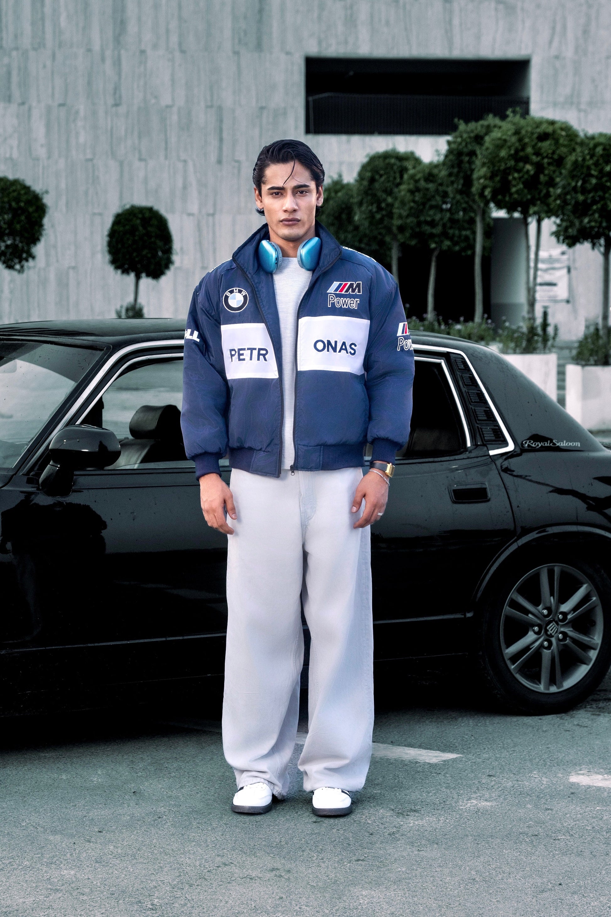 Koti Apparels BMW racing jacket Pakistan with embroidered motorsport logos worn by a man standing next to a black car.