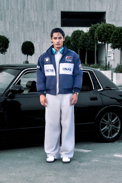 Koti Apparels BMW racing jacket Pakistan with embroidered motorsport logos worn by a man standing next to a black car.