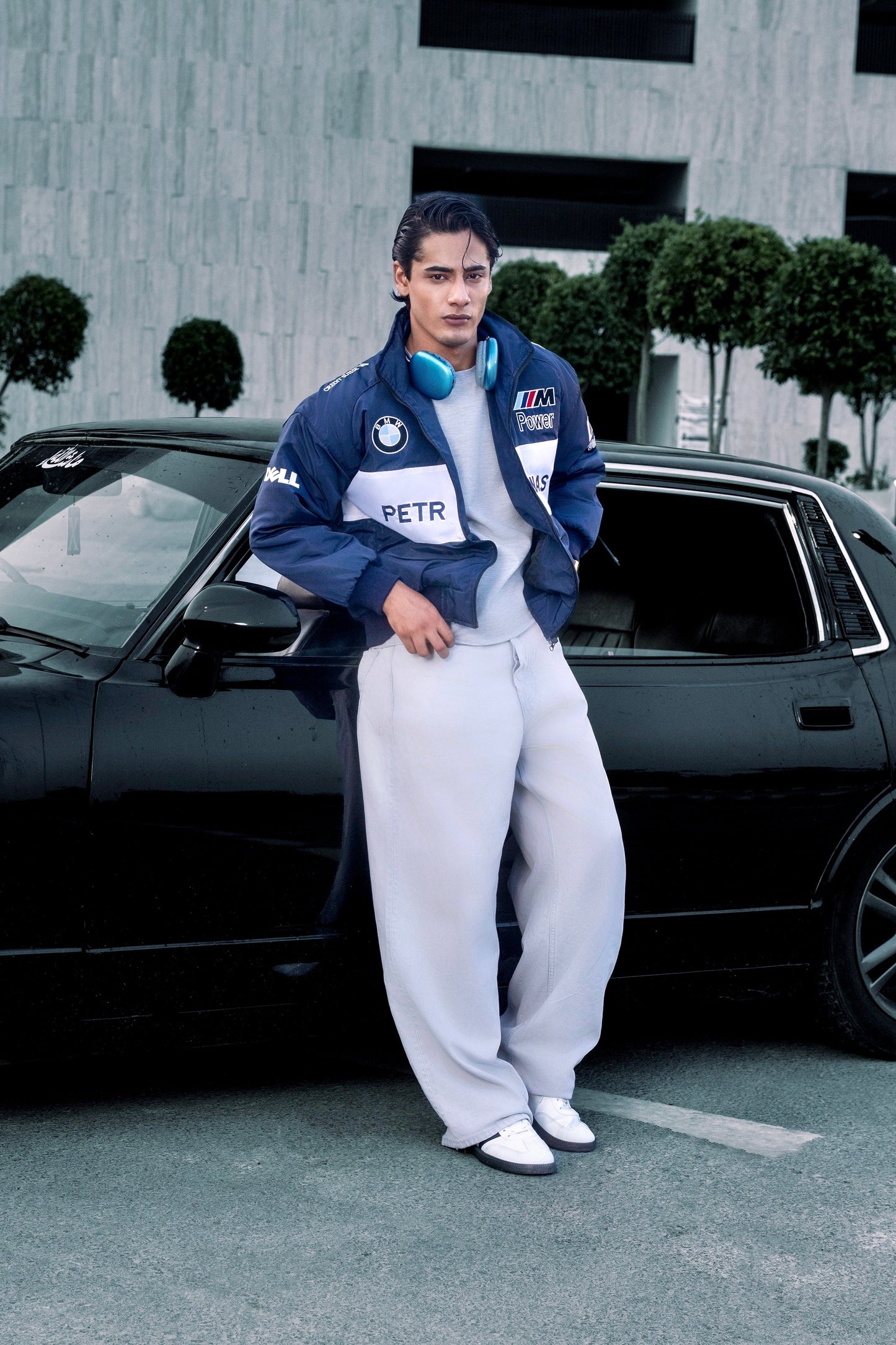 Model wearing a Koti Apparels BMW racing jacket with embroidered logos, standing by a black car outdoors.