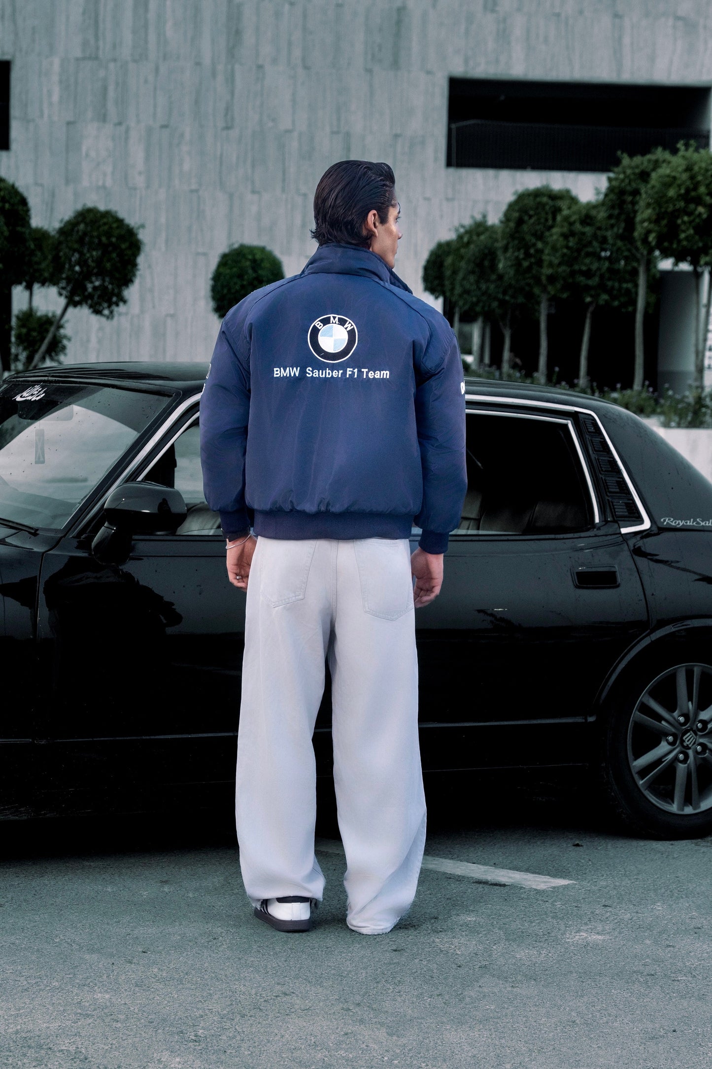 Man wearing a Koti Apparels navy blue BMW racing jacket with embroidered logos, standing by a black car.