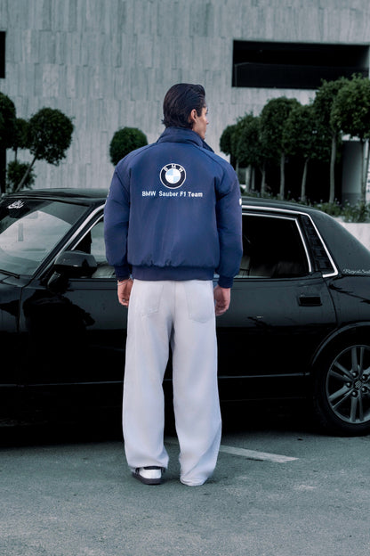 Man wearing a Koti Apparels navy blue BMW racing jacket with embroidered logos, standing by a black car.