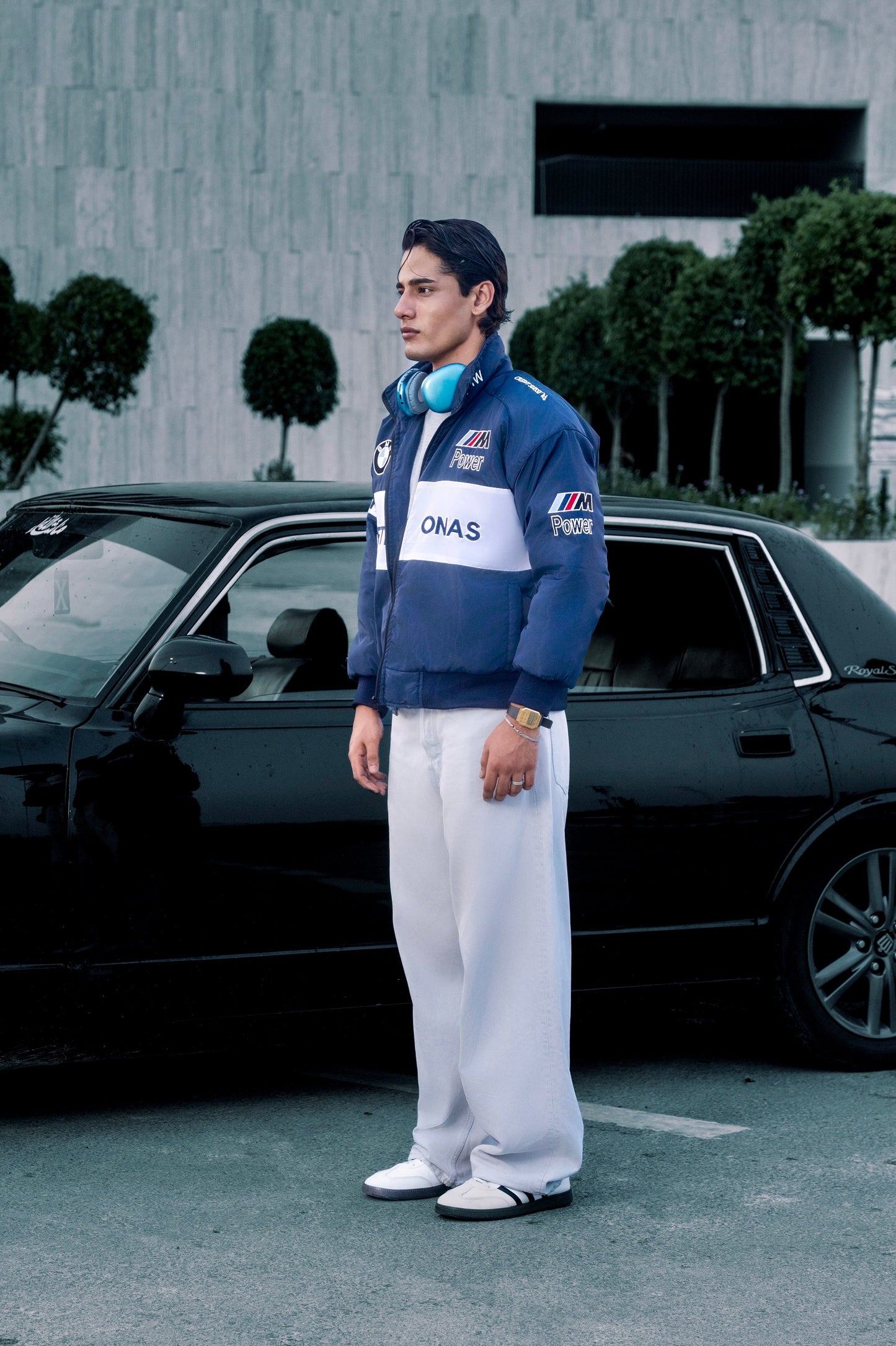 Koti Apparels BMW racing jacket in blue with embroidered motorsport logos worn by a man standing by a black car