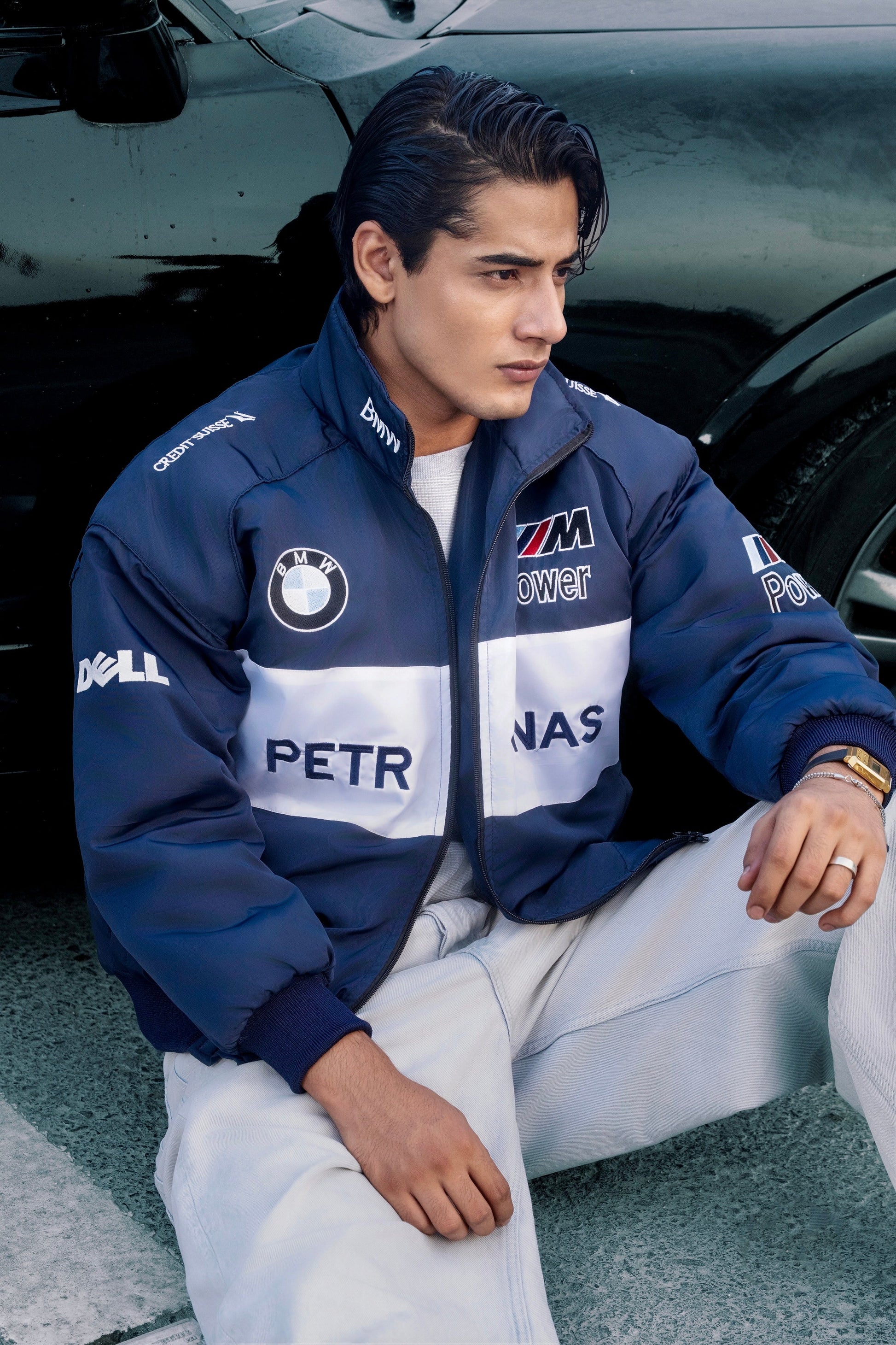 Koti Apparels BMW racing jacket in navy and white with embroidered motorsport logos worn by a man near a black car