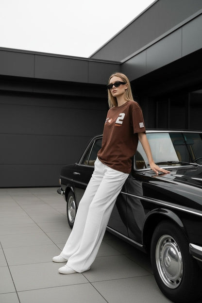 Koti Apparels brown cotton t-shirt with graphic patches and bold numbering, worn by a woman leaning on a vintage car.