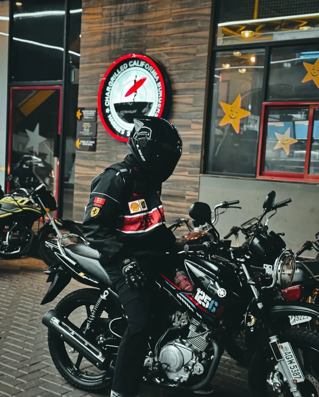 Koti Apparels black parachute fabric F1 Ferrari jacket with embroidered logos and red/white stripe detail. Model wears helmet and sits on a motorcycle.