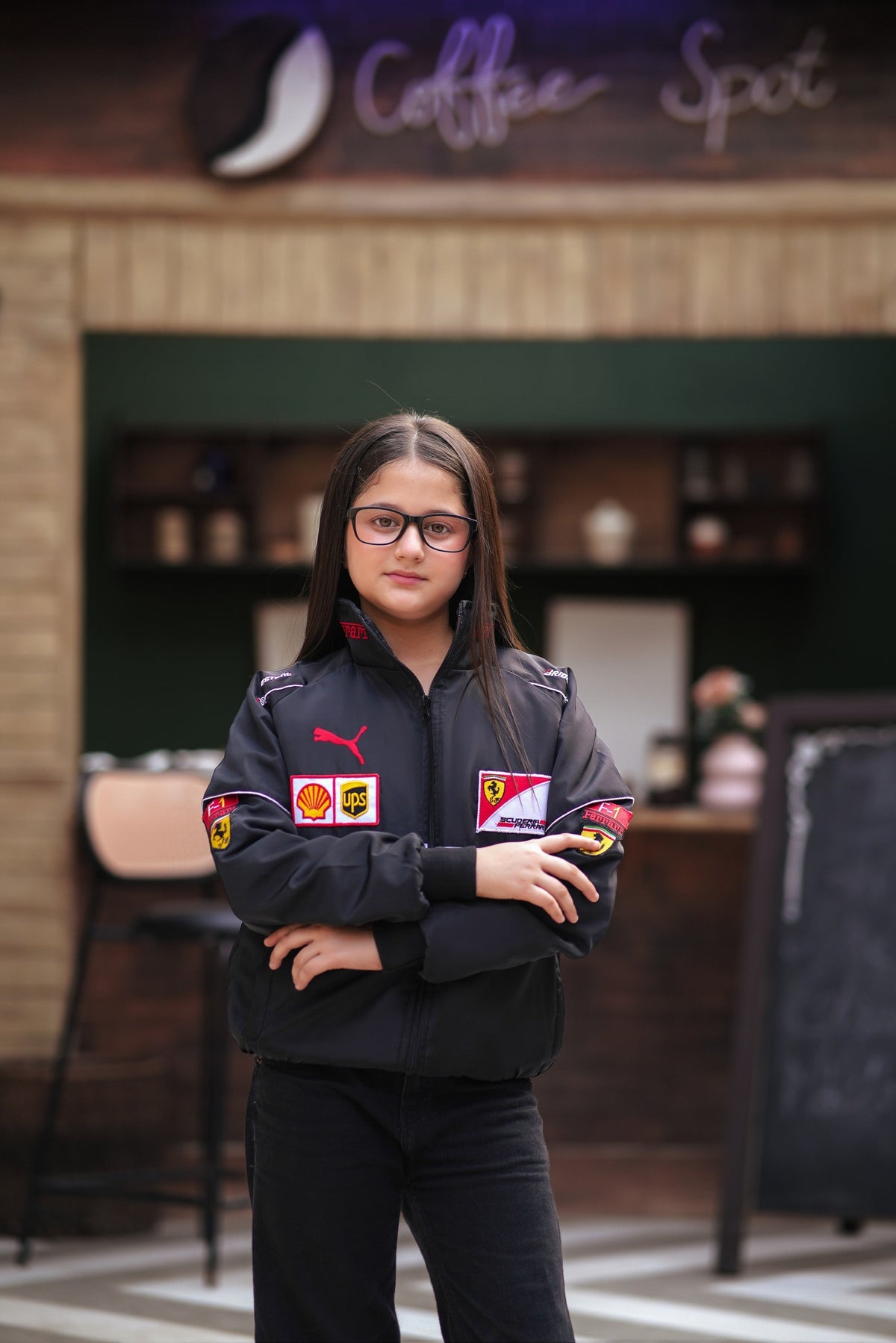 A young girl wearing a black Koti Apparels Ferrari racing jacket with red and white sponsor logos and Puma branding.