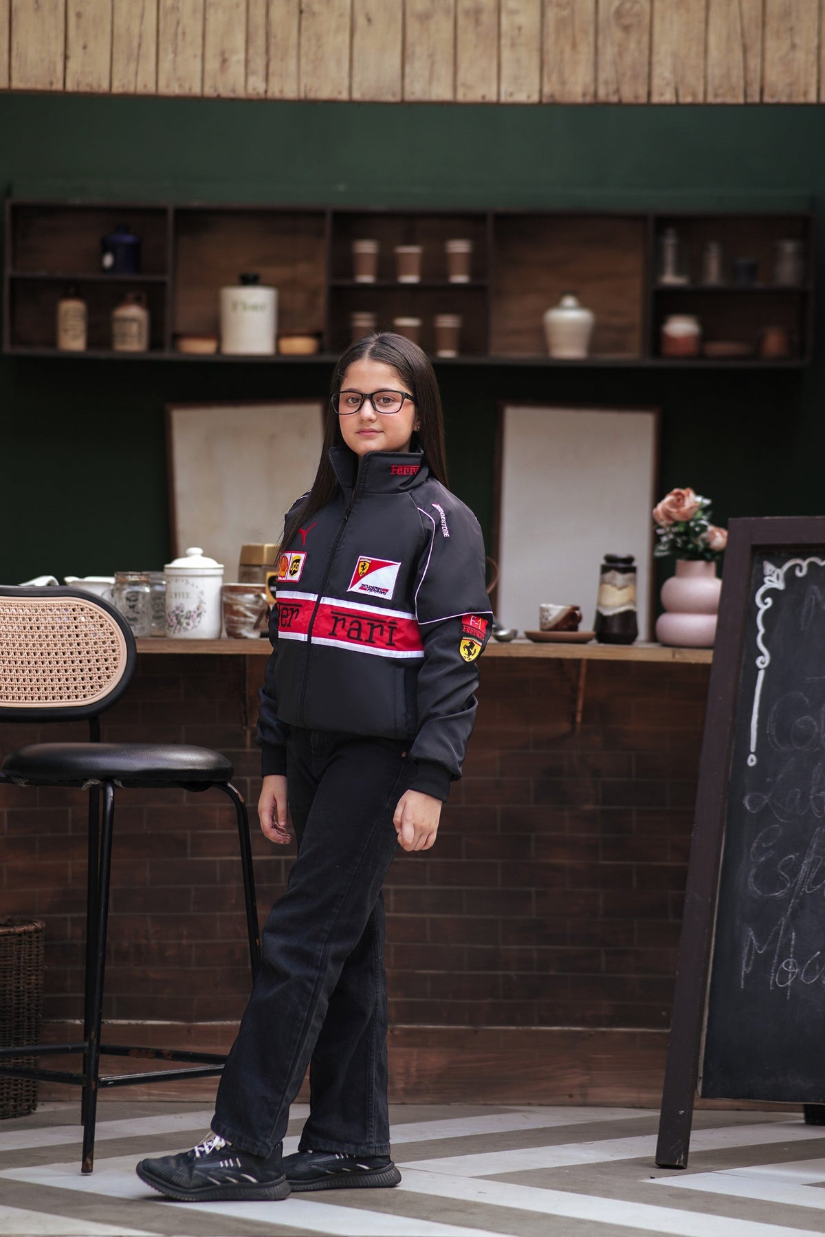 Koti Apparels black Ferrari racing jacket for kids with red and white accents, worn by a girl in a cafe setting.