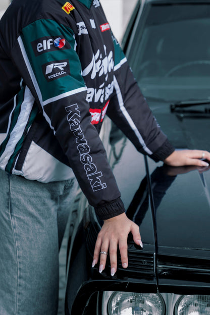 Koti Apparels Kawasaki racing jacket in black and green polyester with sponsor logos, worn by a model near a car.