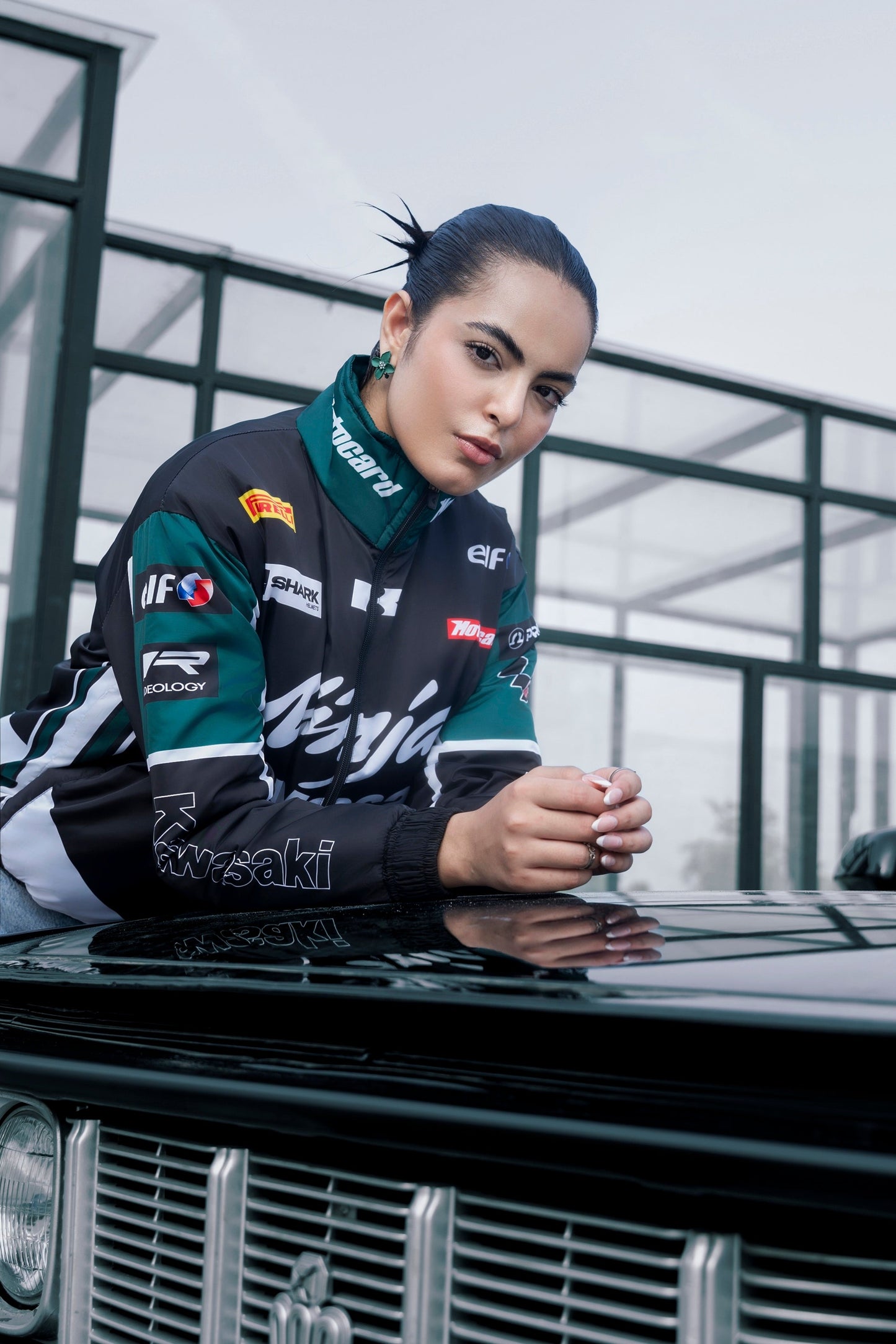 Woman wearing a Koti Apparels Kawasaki racing jacket in black and green with sponsor logos, leaning on a car hood.