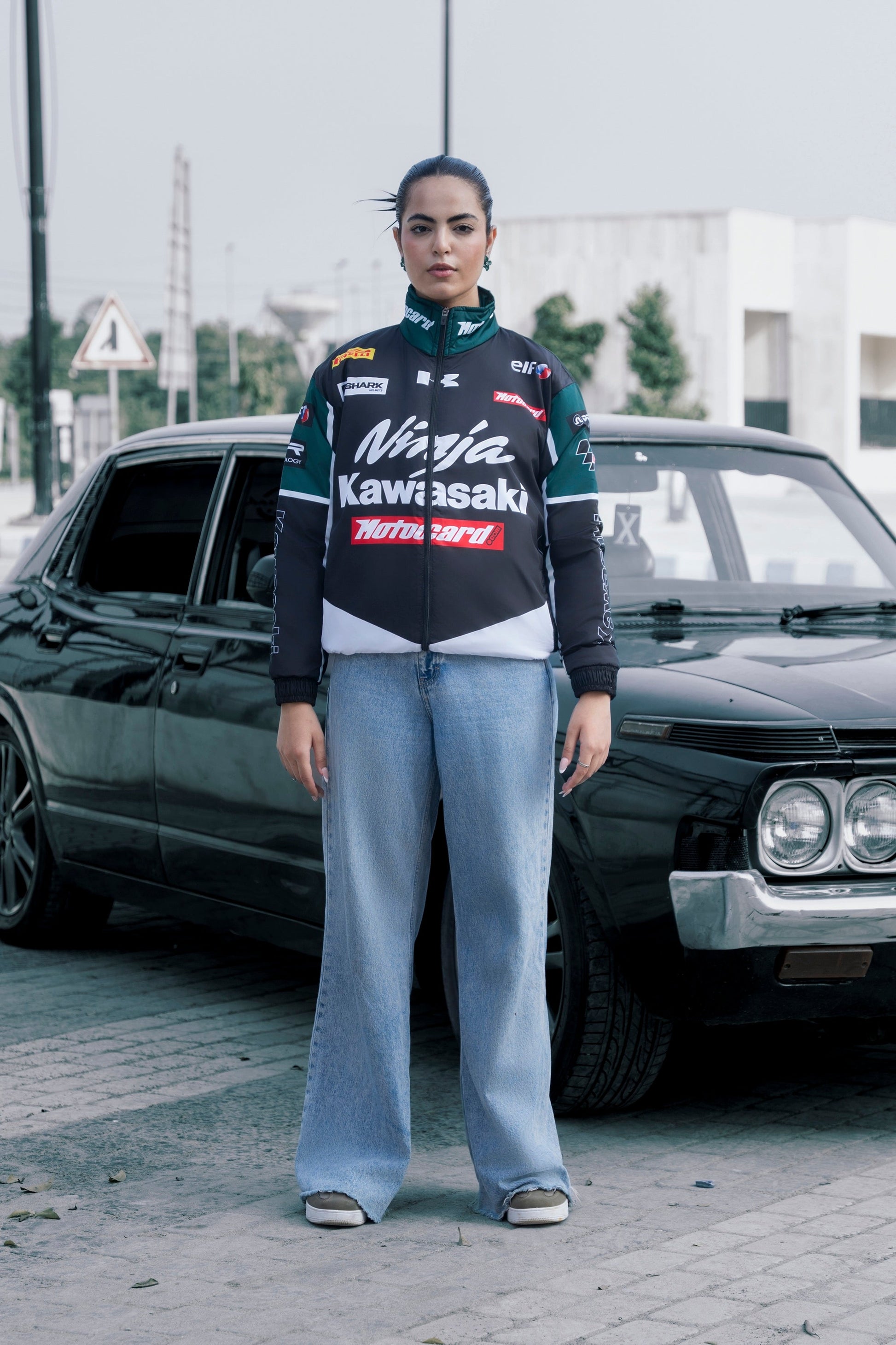 Model wearing a Koti Apparels Kawasaki racing jacket in black and green with sponsor logos, paired with blue jeans.