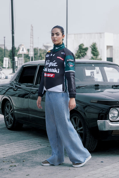Model wearing a Koti Apparels Kawasaki racing jacket in black and green with sponsor logos, paired with wide-leg jeans