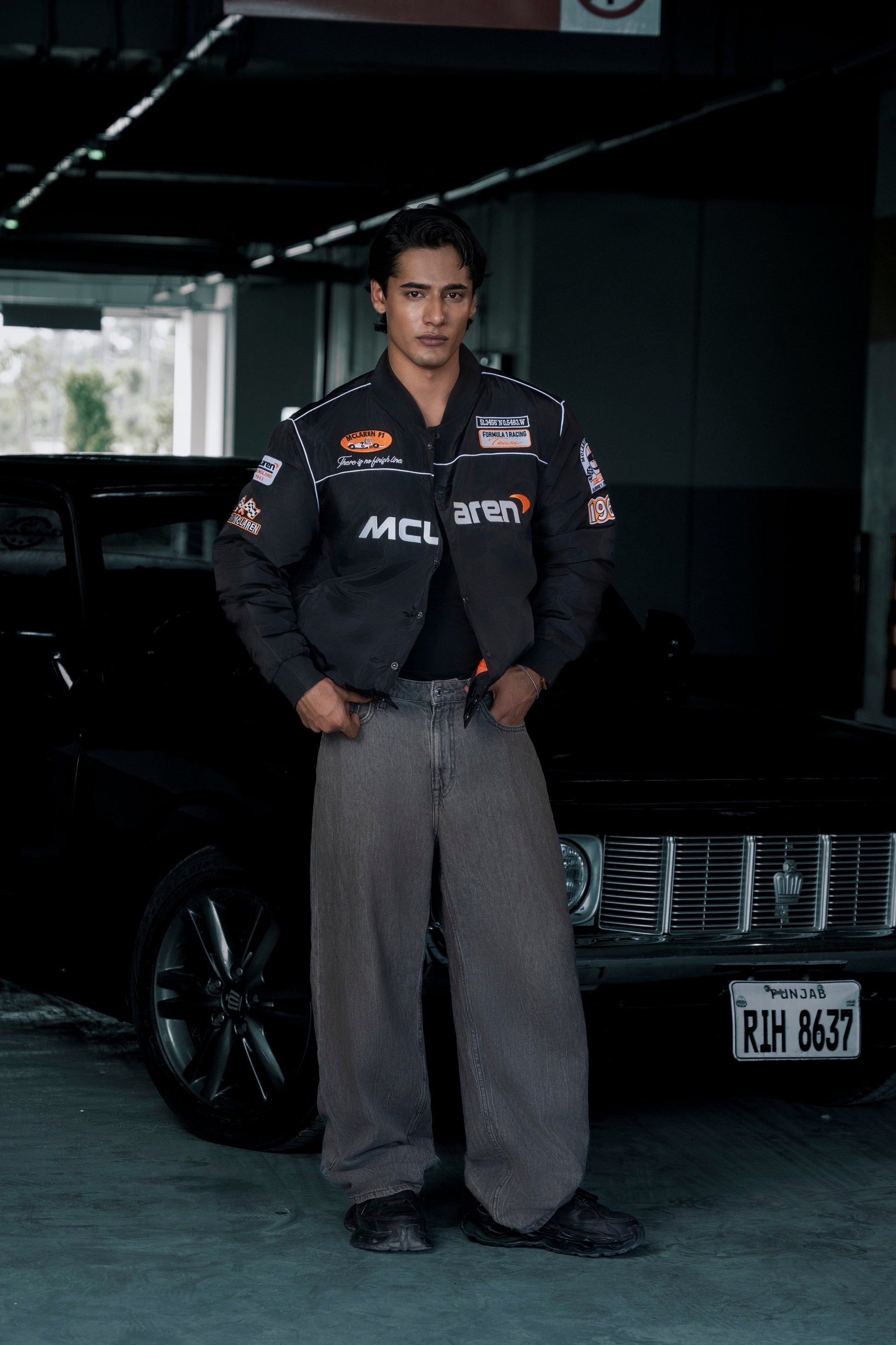 Koti Apparels McLaren F1 racing jacket in black with premium needle embroidery, worn by a man standing by a car.