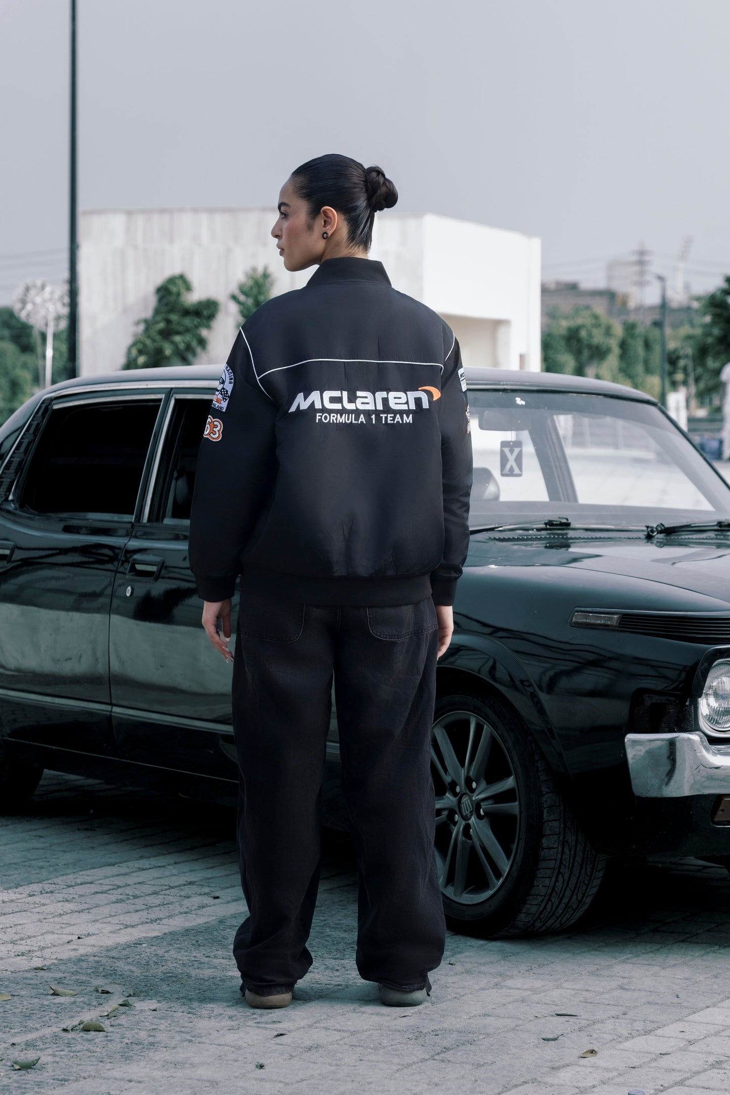 Koti Apparels black McLaren F1 racing jacket with premium embroidery, worn by a model standing by a vintage car.