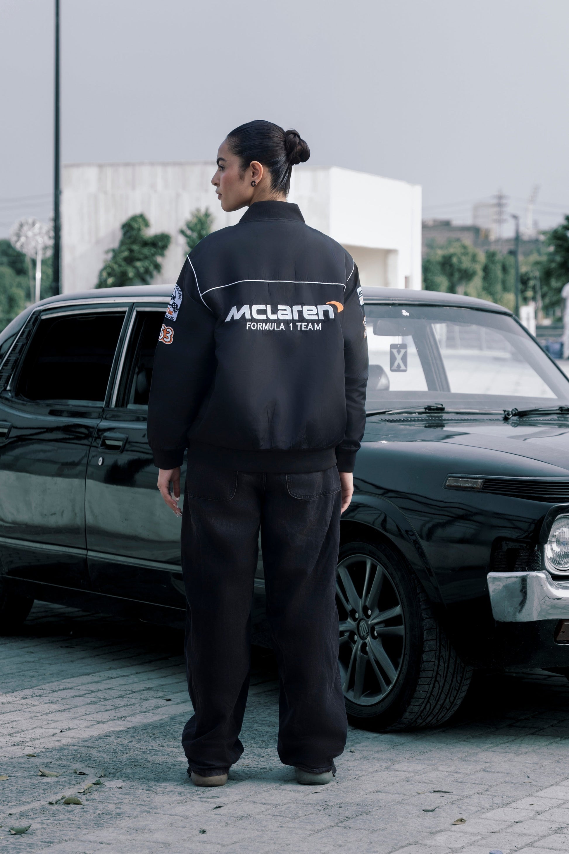Koti Apparels black McLaren F1 racing jacket with premium embroidery, worn by a model standing by a vintage car.