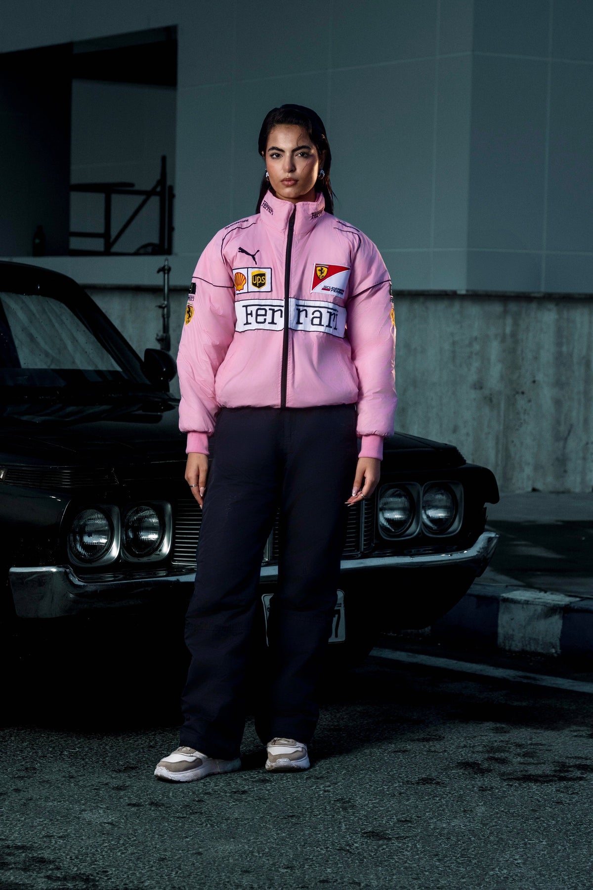 Model wearing a Koti Apparels pink Ferrari racing puffer jacket with embroidered logos, standing by a black car.
