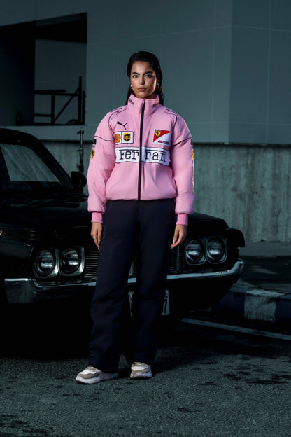 Model wearing a Koti Apparels pink Ferrari racing puffer jacket with embroidered logos, standing by a black car.