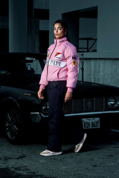 Model wearing a Koti Apparels pink Ferrari racing jacket with embroidered logos, standing by a black vintage car.