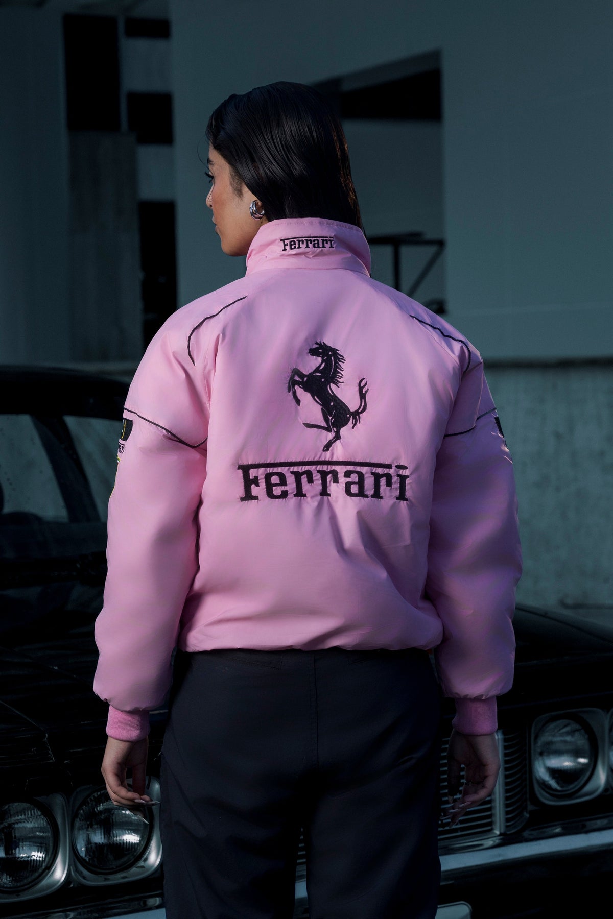 A woman wearing a Koti Apparels pink Ferrari racing jacket with black embroidered logos, viewed from the back.