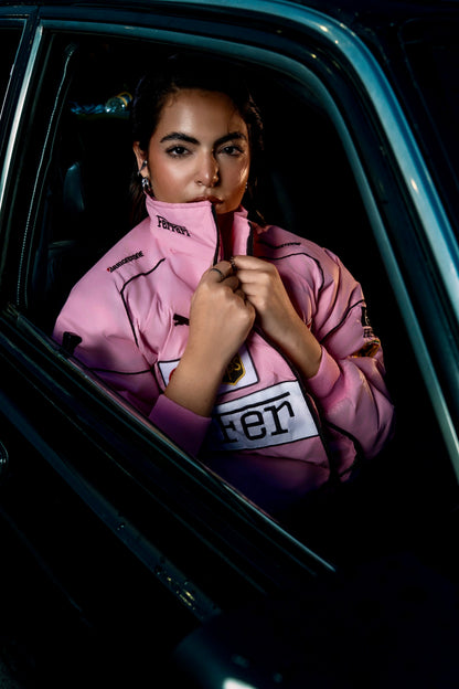 A woman wearing a Koti Apparels pink Ferrari racing jacket with embroidered logos, sitting inside a car.