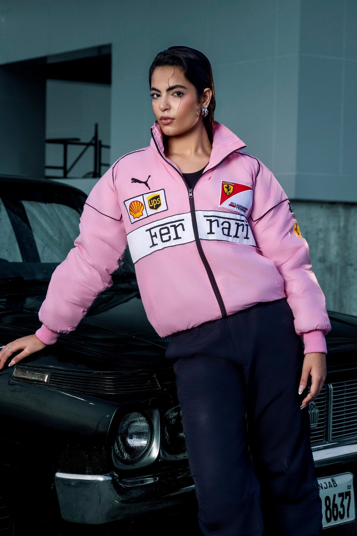 Koti Apparels pink Ferrari racing jacket with embroidered logos worn by a woman leaning against a black car